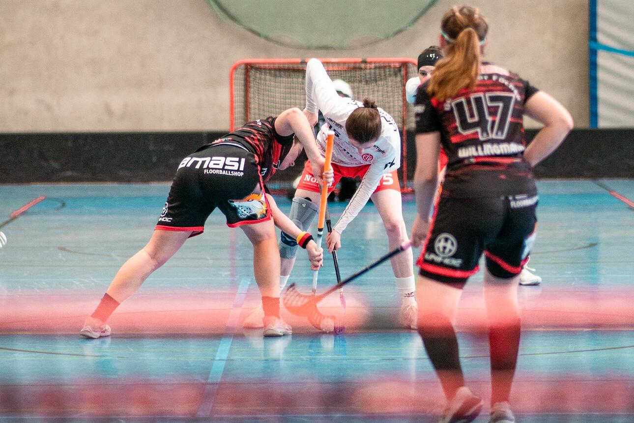 Spielerinnen kämpfen um den Ball in einem Hallenbereich des Floorballspiels mit einem Tor im Hintergrund.
