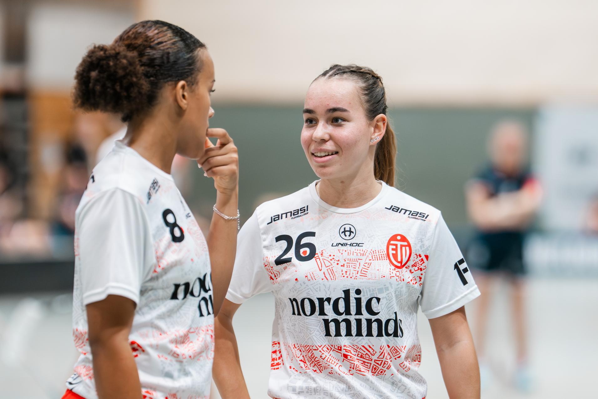 Zwei weibliche Sportlerinnen im Gespräch, tragen hellen Trikots mit roten Hosen und halten ihre Schläger in einer Sporthalle.