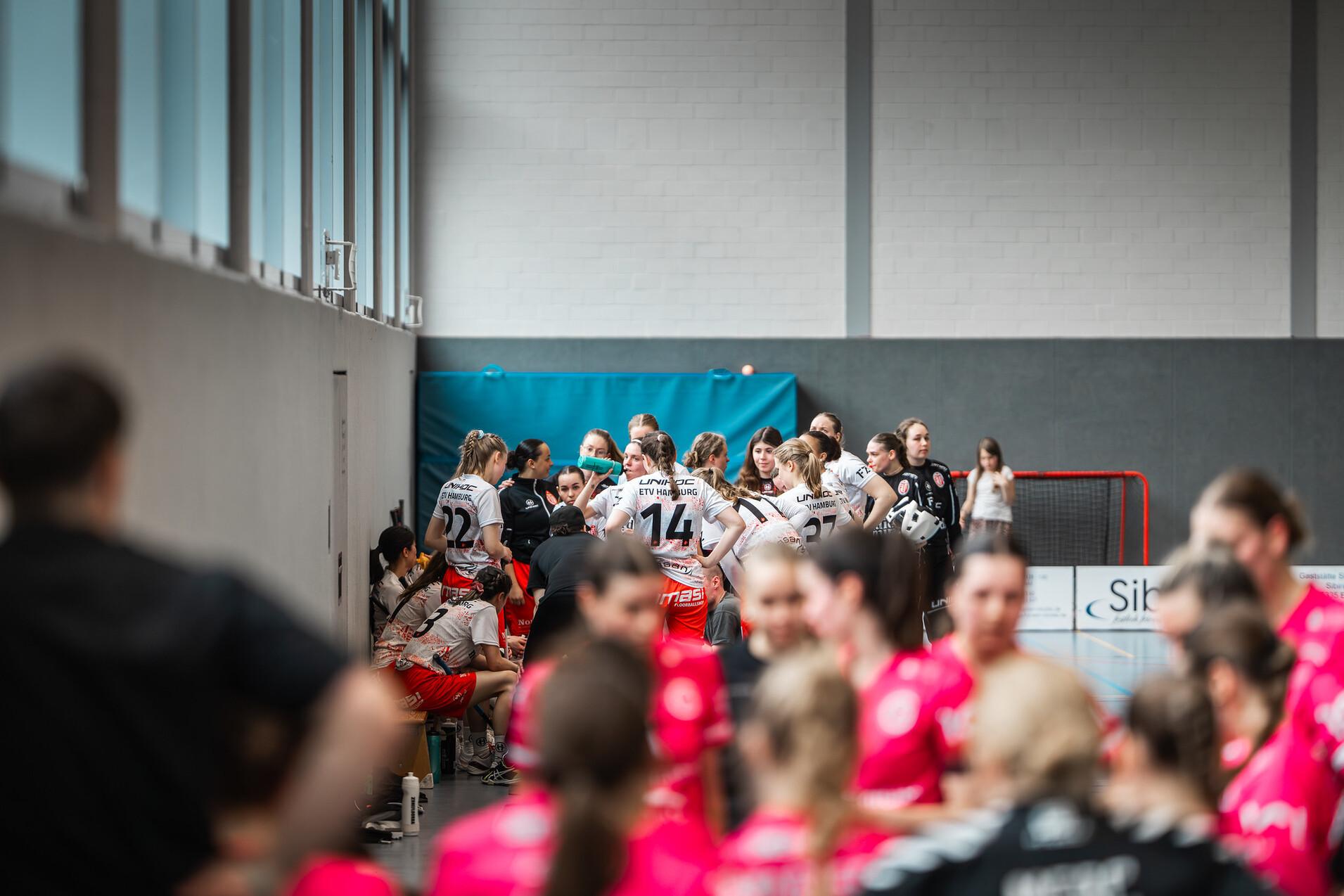 Szenenaufnahme einer Sportmannschaft in einem Meeting, mit Spielerinnen in unterschiedlichen Trikots in einer Halle.