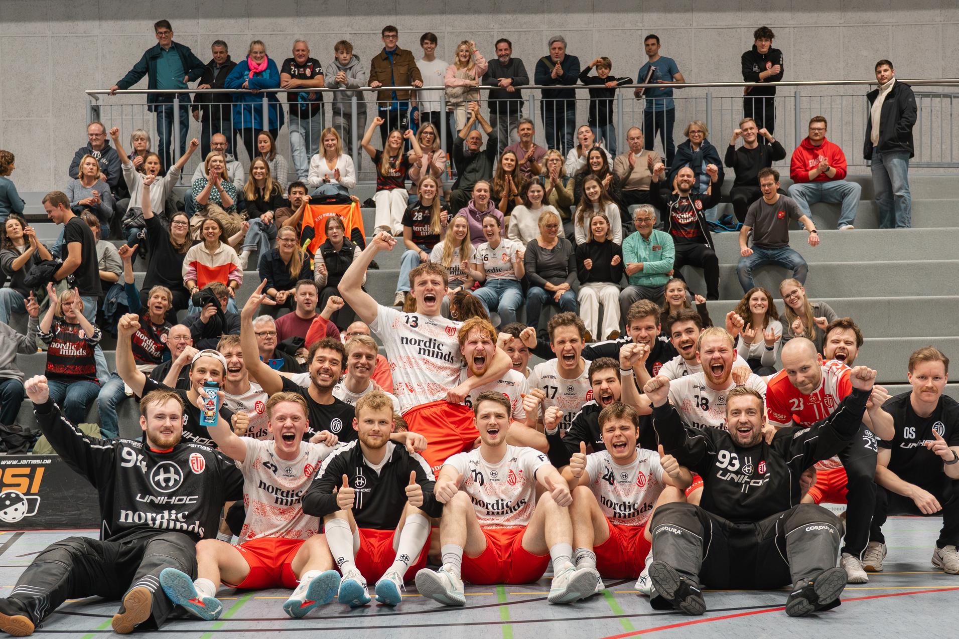 Gruppe von Handballspielern und Fans jubelt gemeinsam in einer Sporthalle, fröhliche Stimmung und emotionale Freude.