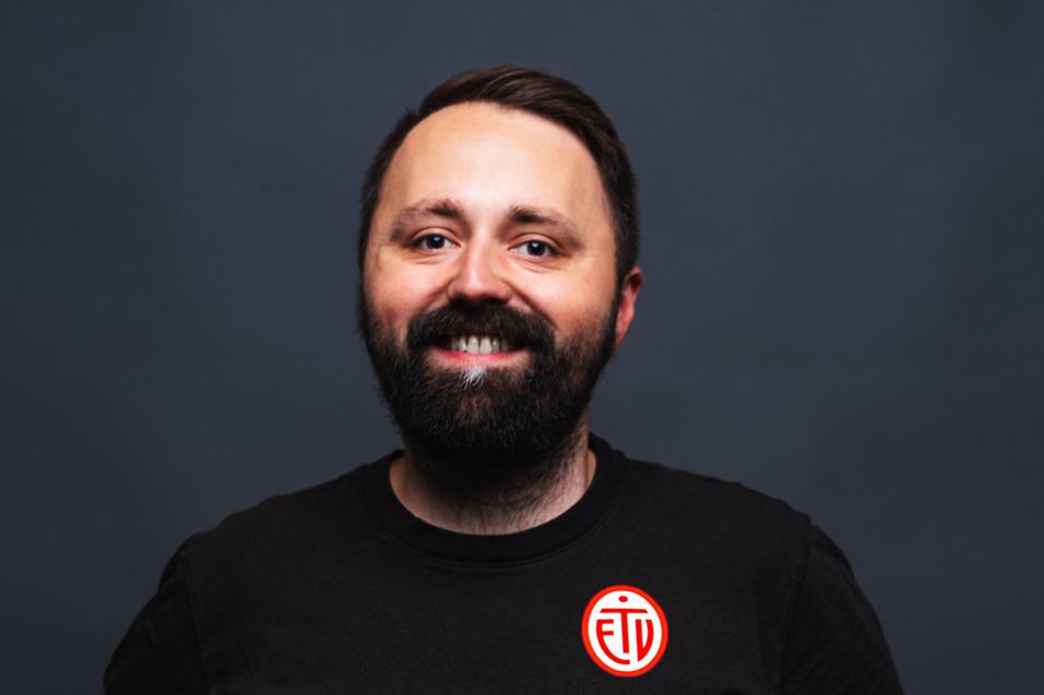 Porträt eines lächelnden Mannes in schwarzem T-Shirt mit Logo, vor einem neutralen, dunklen Hintergrund.