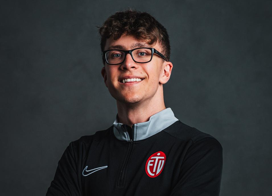 Junger Mann mit Brille in schwarzem Sportpullover, lächelt und hat die Arme vor der Brust verschränkt, vor neutralem Hintergrund.