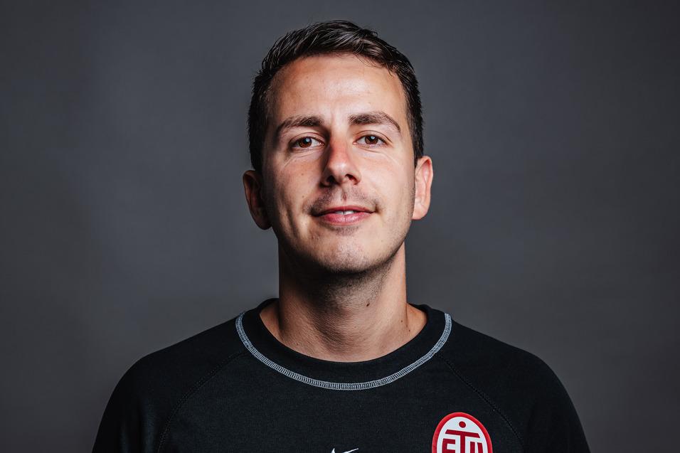 Porträt eines lächelnden Mannes in einem schwarzen Sportshirt mit Logo der Atamimi Group und einem Sportteamsymbol.