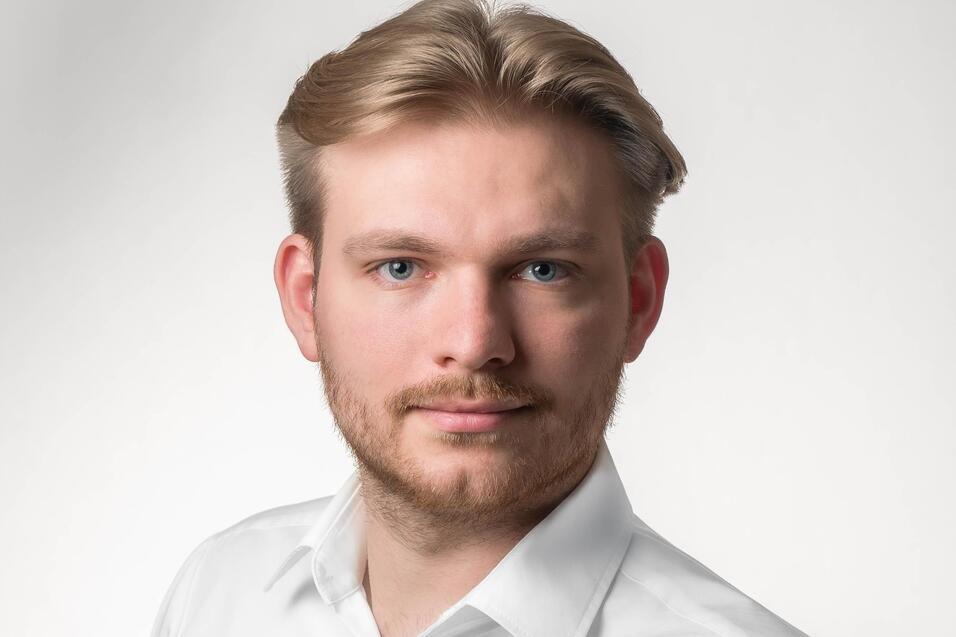 Porträt eines Mannes mit blonden Haaren und Bart, in einem weißen Hemd vor einem hellen Hintergrund.