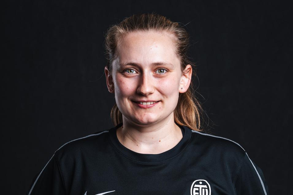 Porträt einer lächelnden Frau in schwarzer Sportkleidung mit Logo, vor einem dunklen Hintergrund.
