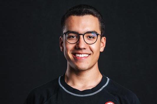 Lächelnder junger Mann mit Brille trägt ein schwarzes T-Shirt mit Logo auf dunklem Hintergrund.