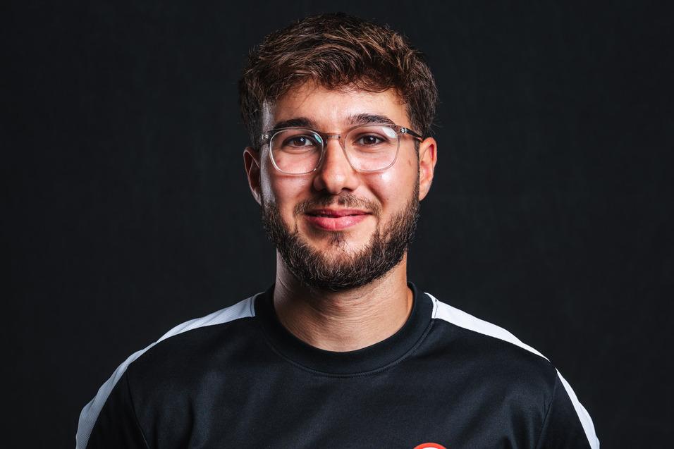 Junger Mann mit Brille in schwarzem Nike-Trikot mit Logo, lächelt vor dunklem Hintergrund.