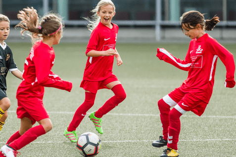 Vier Mädchen in roten Fußballtrikots spielen begeistert auf einem Kunstrasenplatz mit einem Fußball.