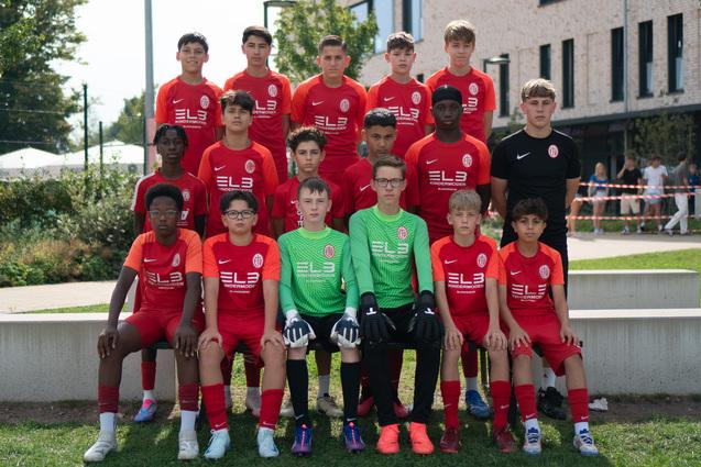 Gruppe von 15 Fußballspielern in roten Trikots, darunter ein Torwart in grüner Ausrüstung, posiert auf einem Platz.