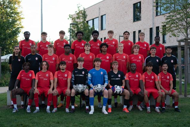 Gruppenfoto von 22 jungen Fußballspielern in roten Trikots, zusammen mit zwei Torhütern in blauen und schwarzen Trikots.