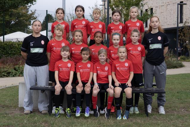 Gruppenfoto einer weiblichen Jugendfußballmannschaft in roten Trikots, auf einer Wiese vor einem Gebäude.