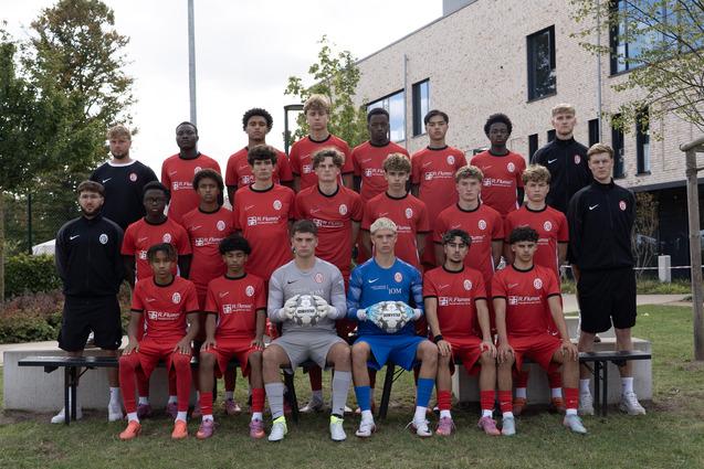 Mannschaftsfoto einer Fußballmannschaft in roten Trikots, sitzend und stehend vor einem modernen Gebäude.