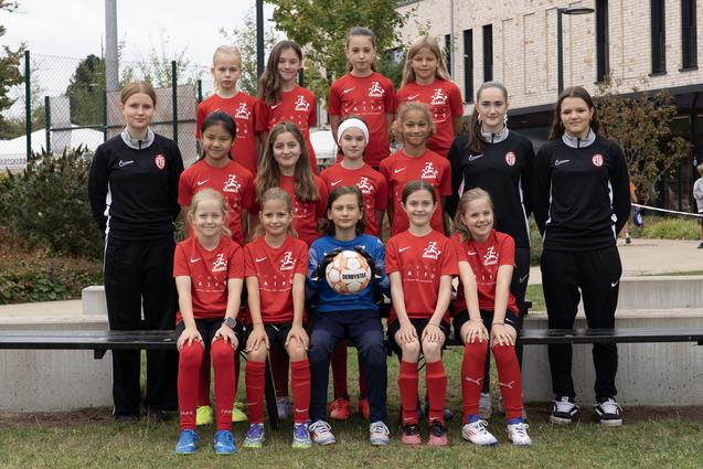 Gruppenfoto eines Mädchenteams in roten Fußballtrikots, sitzend auf einer Bank vor einem modernen Gebäude.