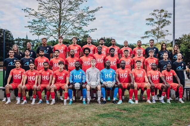 Mannschaftsaufstellung eines Fußballteams in roten Trikots auf Rasen, umgeben von Bäumen und einem wolkigen Himmel.