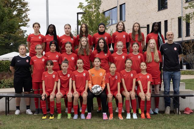 Mannschaftsfoto eines weiblichen Jugendfußballteams in roten Trikots, posing mit einem Fußball auf einer Wiese.