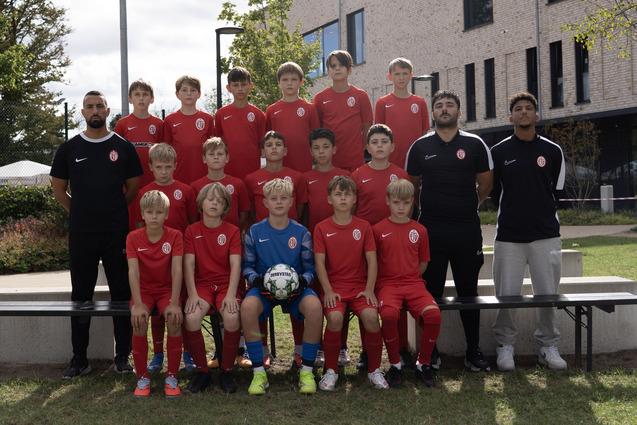 Gruppenfoto einer Jugendfußballmannschaft in roten Trikots, umgeben von Bäumen und einem modernen Gebäude.