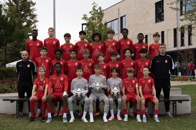 Gruppenbild von einer Jugendfußballmannschaft in roten Trikots, umgeben von Bäumen und modernem Gebäude.