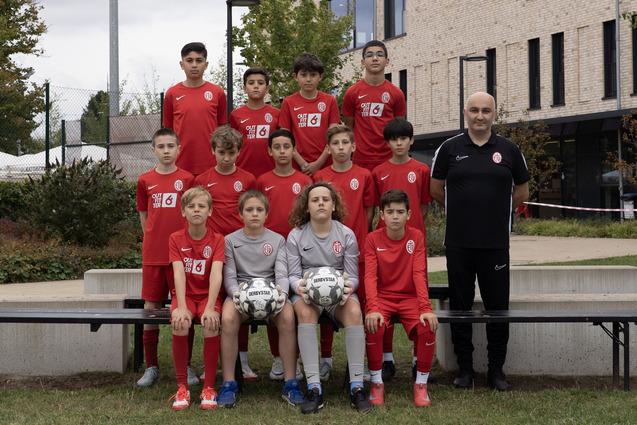 Gruppenfoto einer Jugendfußballmannschaft in roten Trikots, posierend auf einer Bank vor einem modernen Gebäude.
