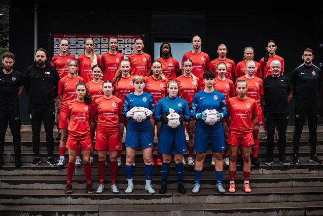 Mannschaftsbild einer Frauenfußballmannschaft in roten Trikots, auf einer Treppe vor einer Sporteinrichtung stehend.