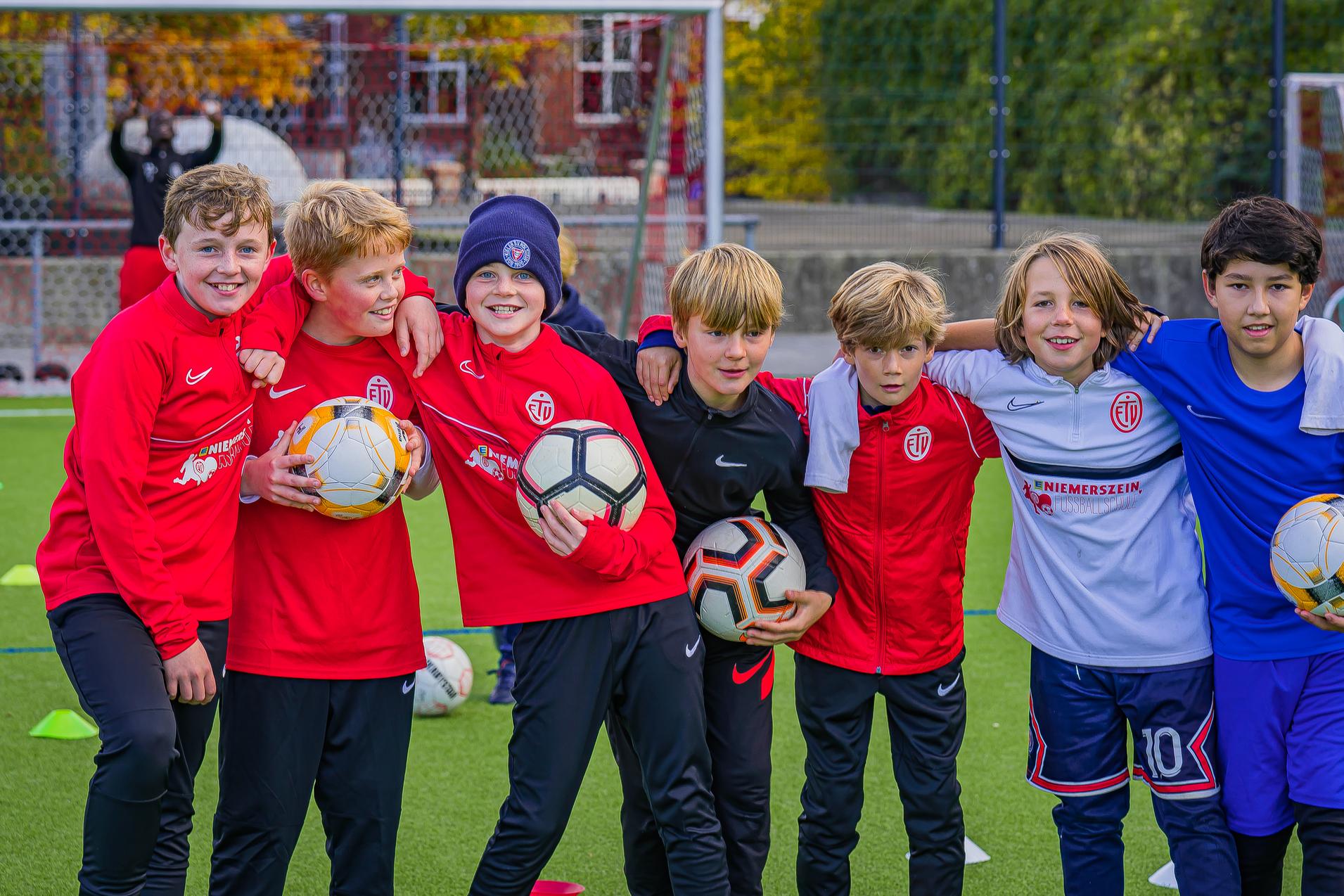 Gruppe von sieben Jungen in Sportkleidung mit Fußbällen auf einem Fußballplatz, lächelnd und zum Foto posierend.