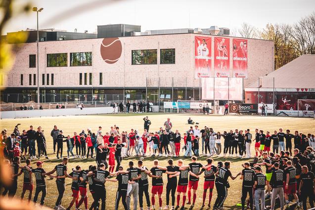 Gruppierung von Spielern und Zuschauern auf einem Sportplatz, um einen Kreis mit dem Vereinsgebäude im Hintergrund zu bilden.