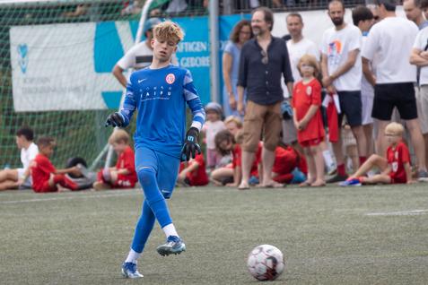Junger Torwart in blauer Sportausrüstung tritt einen Ball auf einem Rasenspielfeld, Zuschauer im Hintergrund.