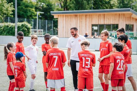 Gruppe von Kindern in roten Fußballtrikots versammelt sich um einen Trainer auf einem Sportplatz.