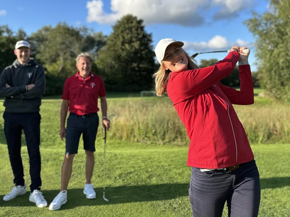 Frau schlägt mit einem Golfschläger auf einer Golfwiese, während zwei Männer im Hintergrund zuschauen.