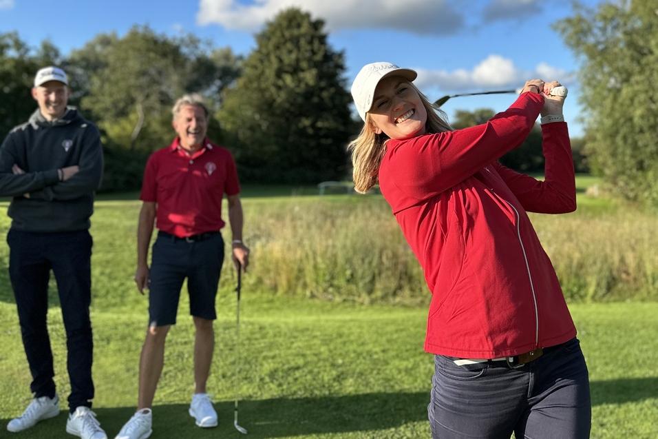 Frau schlägt mit einem Golfschläger auf einer Golfwiese, während zwei Männer im Hintergrund zuschauen.