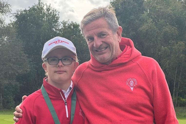 Zwei Personen in roter Sportbekleidung stehen auf einem Golfplatz, einer trägt eine Medaille und beide lächeln.