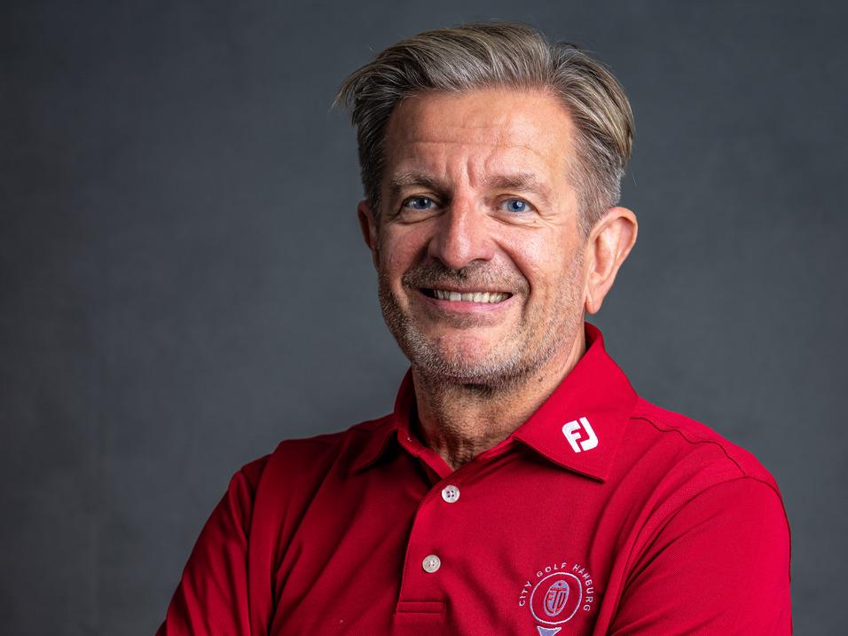 Lächelnder Mann in rotem Poloshirt, mit verschränkten Armen, vor hellem Hintergrund.