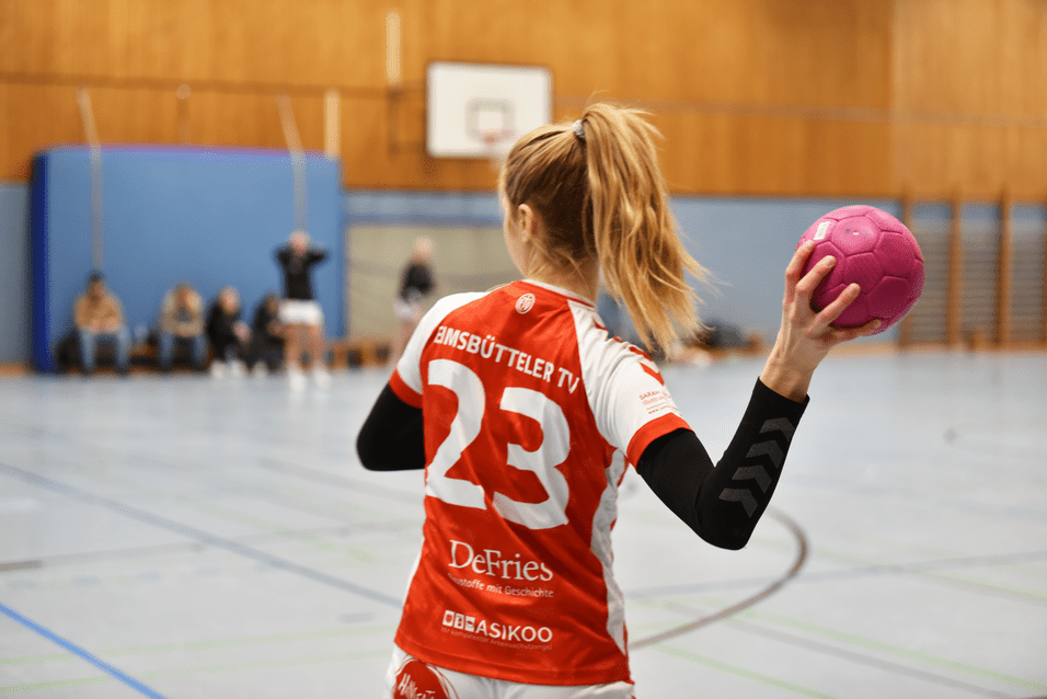 Spielerin des Bimsbütteler TV mit einer pinken Handball im Hallensport, bereit zum Wurf. Rückansicht, Trikotnummer 23.