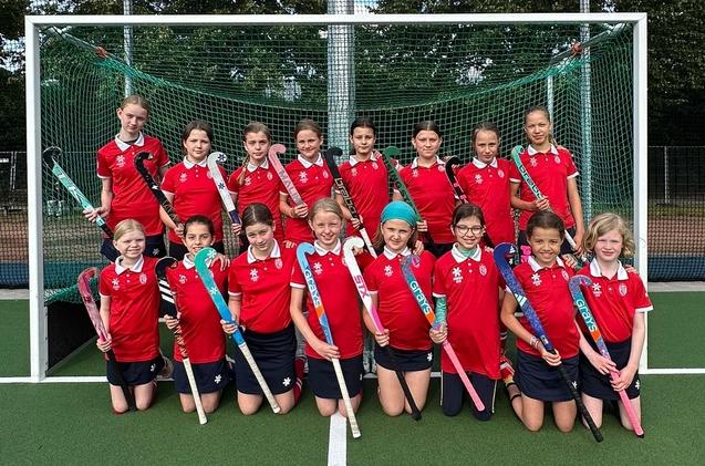 Mädchen-Hockeymannschaft in roten Trikots posiert vor dem Tor mit Hockeyschlägern auf einem Kunstrasenplatz.