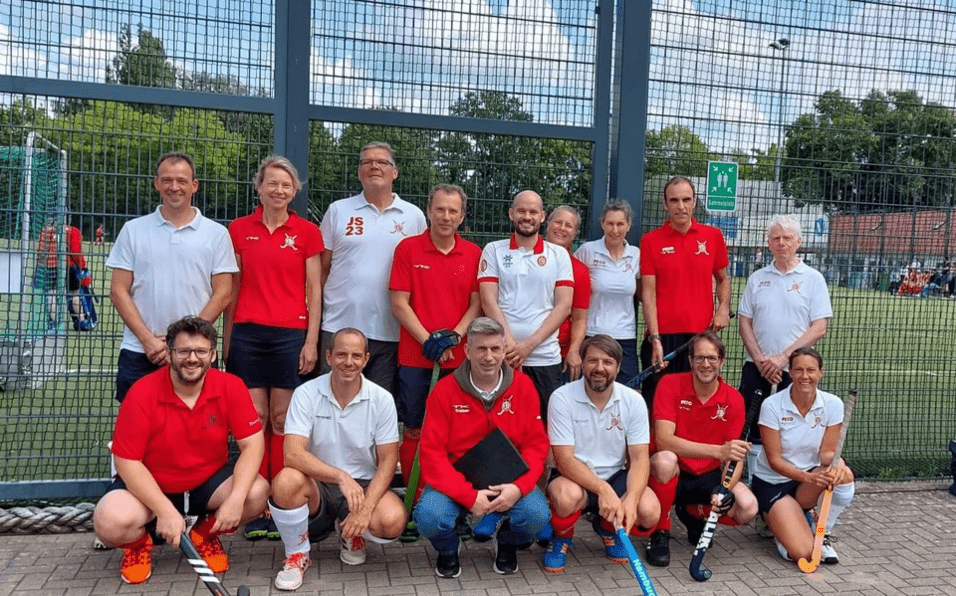 Gruppenfoto von Hockeyspielern in roten und weißen Trikots, stehend und sitzend vor einem Sportplatz mit Maschendrahtzaun.