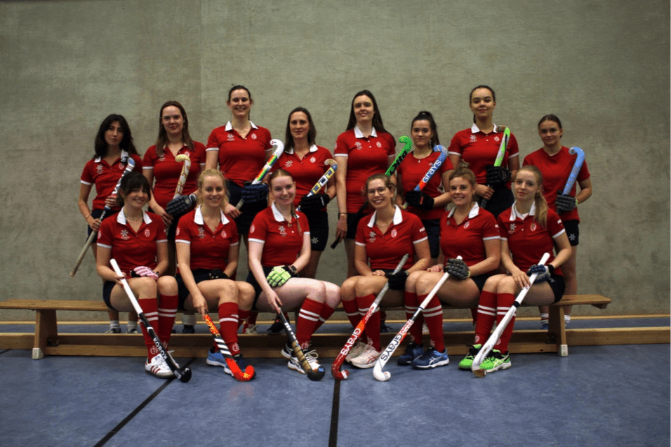Gruppenfoto eines Hockeyteams in roten Trikots mit Schlägern, auf einer Holzbank in einer Sporthalle.