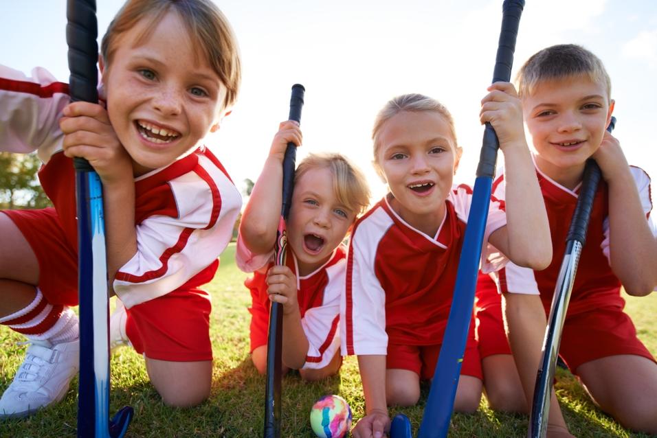 Vier Kinder in roten Sportanzügen lächeln und posieren mit Schlägern auf einer grünen Wiese.