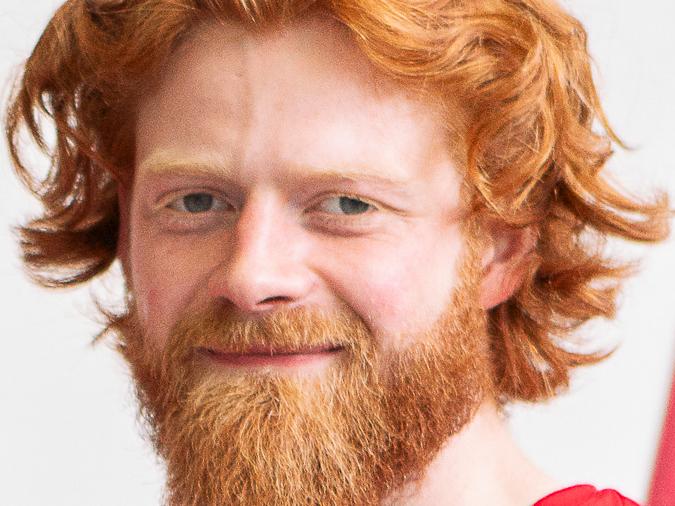 Nahaufnahme eines lächelnden Mannes mit rotem, lockigem Haar und Bart, in einem roten T-Shirt vor neutralem Hintergrund.