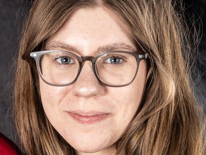 Porträt einer Frau mit langen, braunen Haaren und Brille, vor einem dunklen Hintergrund. Sie lächelt leicht.