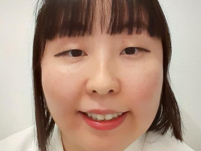 Porträt einer Frau mit kurzen, dunklen Haaren und einem freundlichen Lächeln, trägt einen weißen Kimono. Hintergrund neutral.