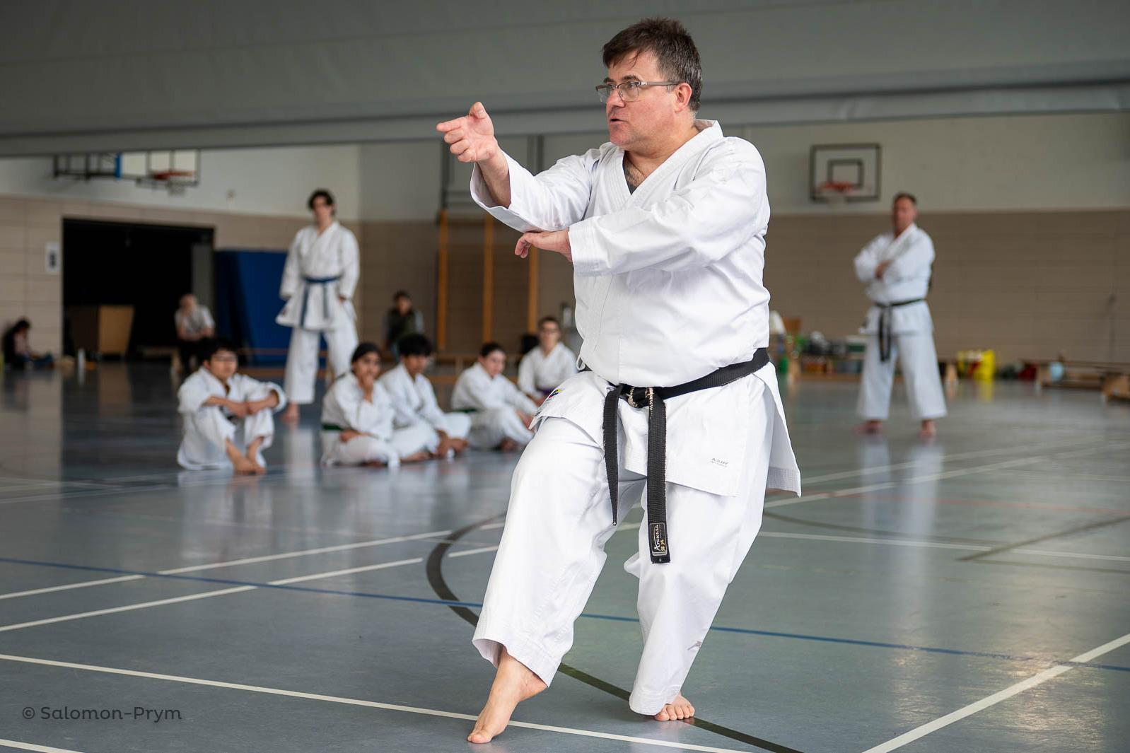 Ein Karateka in Weiß führt einen Angriff im Dojo aus, während andere Trainingsteilnehmer zuschauen.