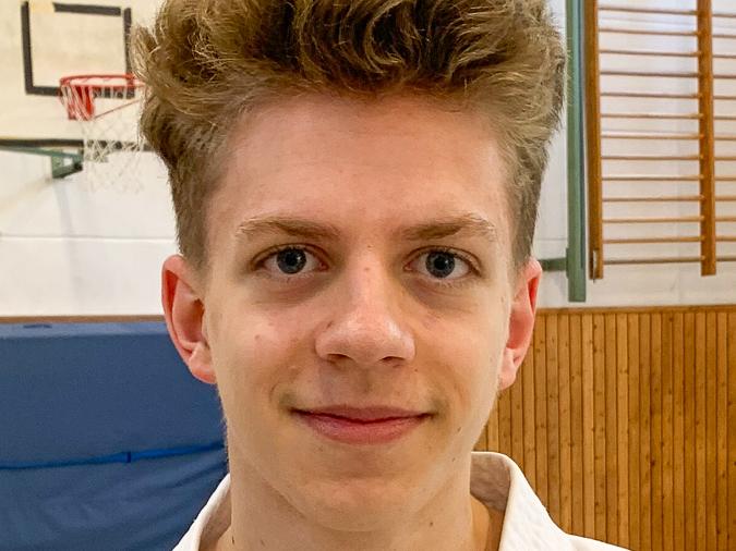 Porträt eines jungen Mannes in einem weißen Karateanzug, aufgenommen in einer Sporthalle mit Holzverkleidung.