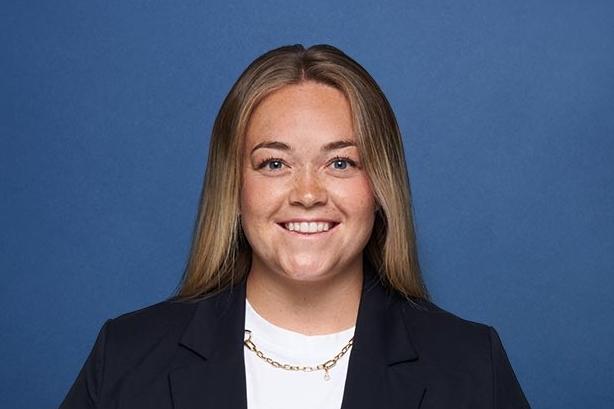 Portrait einer lächelnden Frau mit langen Haaren, in einem dunklen Blazer und weißem Oberteil, vor blauem Hintergrund.