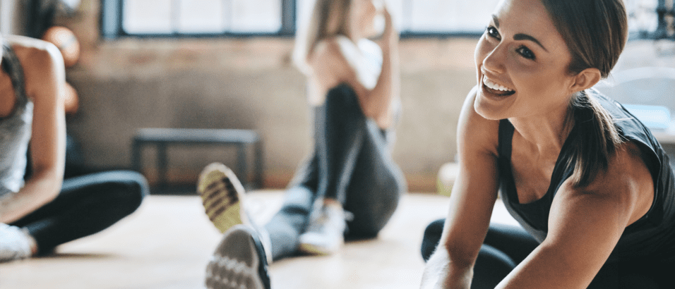 Lächelnde Frau beim Dehnen in einem Fitnessraum mit anderen trainierenden Personen im Hintergrund.