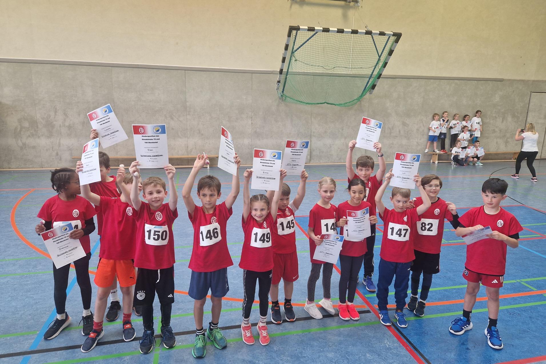 Gruppe von Kindern in roten Shirts hält Urkunden in der Sporthalle, ansässige Torwand im Hintergrund.