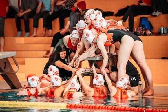 Wasserballmannschaft feiert am Beckenrand, während Trainerin sie anfeuert und Spielerinnen sich um einen Podest versammeln.