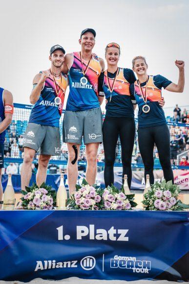 Zwei Männer und zwei Frauen mit Medaillen feiern auf dem Treppchen den ersten Platz bei einem Beachvolleyballturnier.