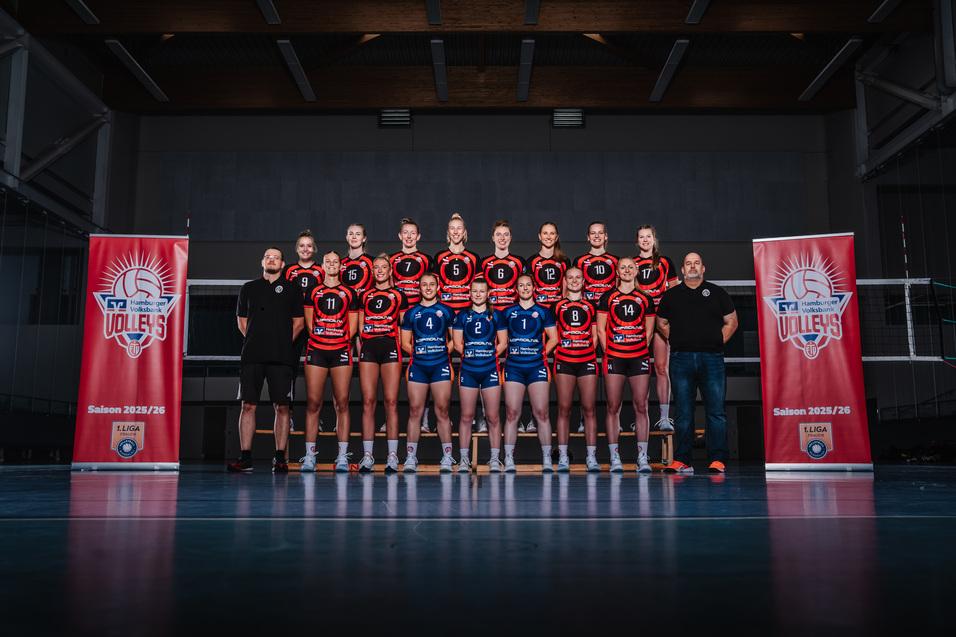 Teamfoto einer Volleyballmannschaft mit Spielerinnen in roten und blauen Trikots, umgeben von Sponsorenbannern.
