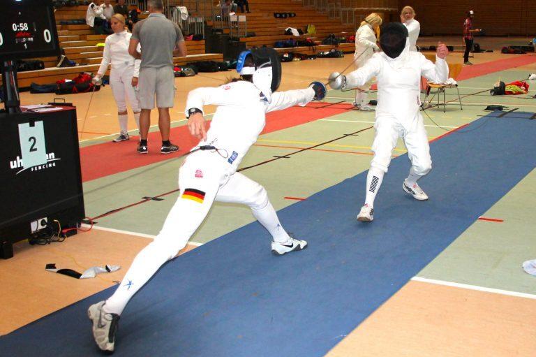 Zwei Fencer in Schutzkleidung führen einen Wettkampf auf einer fechtspezifischen Fläche durch.