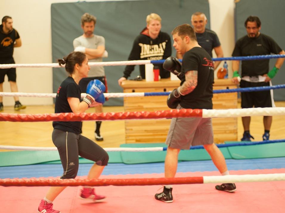Boxtraining in einem Studio: Eine Frau und ein Mann im Ring, umgeben von Zuschauern. Beide tragen Boxhandschuhe.