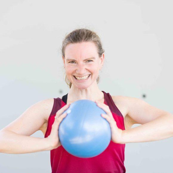 Frau in rot-schwarzem Sporttop lächelt und hält einen blauen Medizinball in beiden Händen.